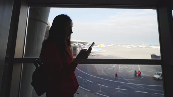 Businesswoman at Airport with Phone in Hands Against the Large Panoramic Window with Parking