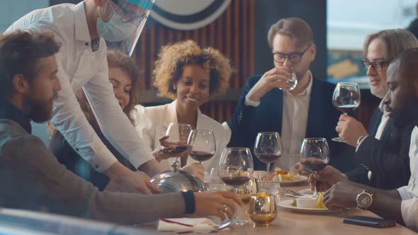 Waiter in Protective Mask Serving Food for Diverse Friends Dining in Modern Restaurant