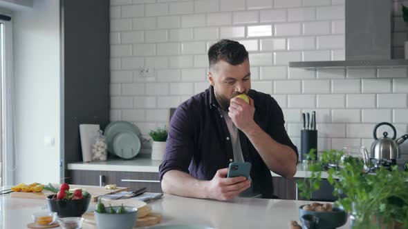 Young Man with Smartphone in the Kitchen searching Online Food Recipe and eating an Apple