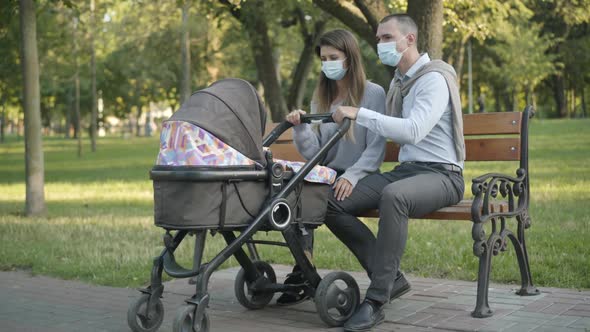 Happy Young Couple of Parents in Covid-19 Face Masks Rocking Baby Stroller and Talking. Wide Shot of
