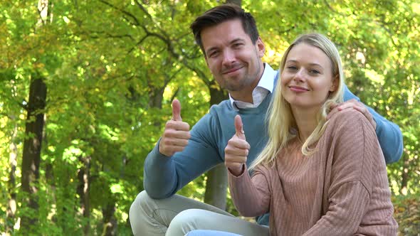 A Young Couple Sits in a Park on a Sunny Day and Shows a Thumb Up To the Camera with a Smile