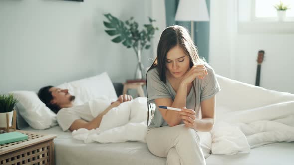 Nervous Girl Sitting on Bed Holding Pregnancy Test While Guy Sleeping in Bedroom