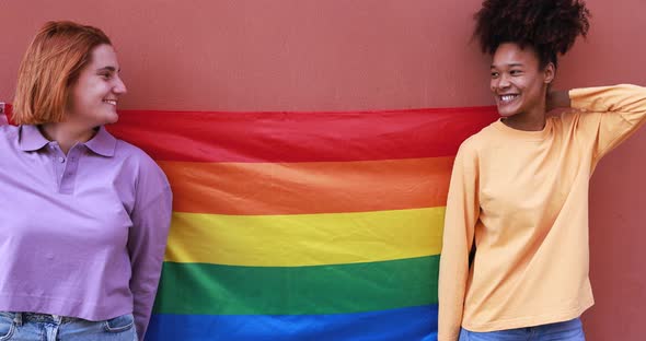 Young multiracial lesbian couple holding LGBT rainbow flag outdoor - Homosexual love concept