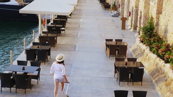 Back View of Young Woman in Summer Clothes and Sun Hat, Walks with Travel Luggage Along a Deserted