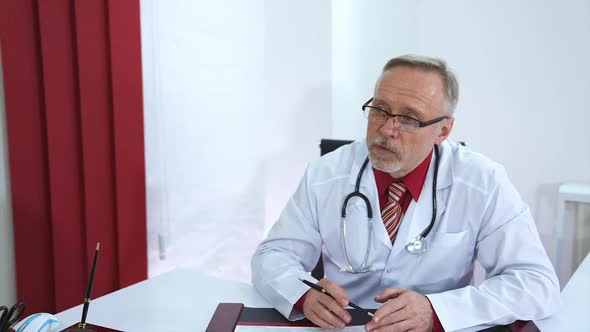 Professional medic sits at table and speaks to a patient. Senior doctor in glasses and grey beard. 