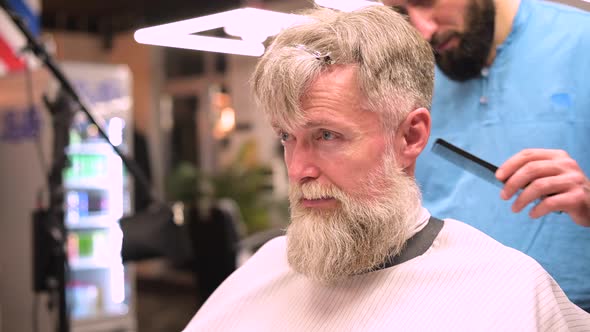 Gray-haired adult bearded man gets a haircut from a barber