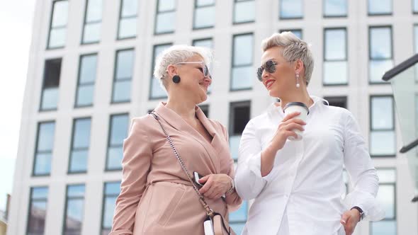 Two Businesswomen Standing Outside with Coffee with Buildings on Background