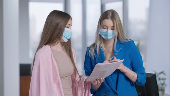 Two Young Beautiful Confident Women in Covid Face Masks Discussing Project in Office with Blurred