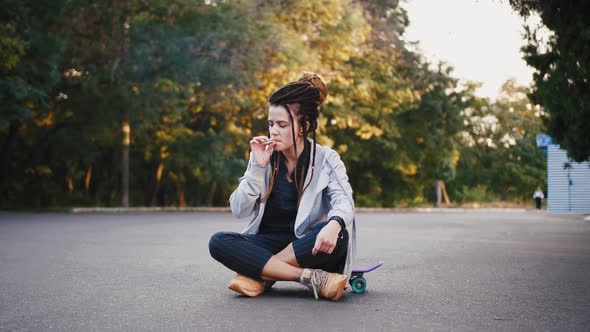 Portrait of Young Pretty Woman with Dreadlocks Sitting on a Skateboard and Smoking Marijuana Joint