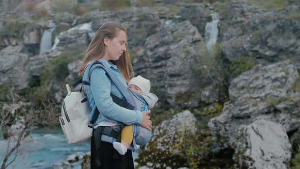 Young mother with her son in ergo backpack travels in nature near the waterfall in spring