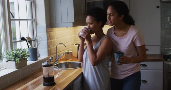 Mixed race lesbian couple hugging and drinking coffee in kitchen