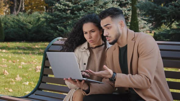 Worried Hispanic Young Couple with Laptop Sit on Bench in Autumn Park Unsuccessful Online Shopping