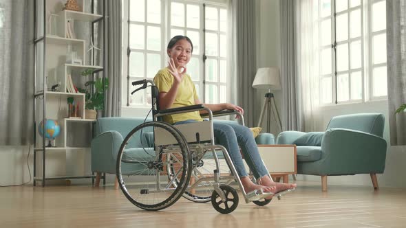 Asian Kid Girl Sitting In A Wheelchair Smiling And Showing Ok Sign In Living Room