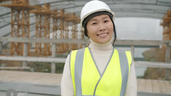 Portrait of Cheerful Asial Lady Wearing Helmet and Protective Vest Smiling in Building Area