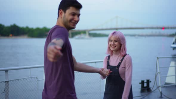 Laughing Beautiful Girlfriend Holding Hand of Cheerful Blurred Boyfriend Pointing at Camera