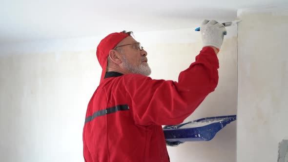 Mature Graybearded Man in a Red Overalls and a Cap Paints a Wall with White Paint