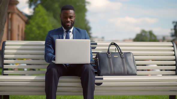 Portrait of Young Attractive African Businessman Sitting on Bench Working on Laptop