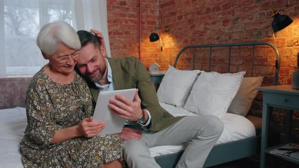 Modern Pensioner Lady Cuddling and Comforting His Middleaged Son While Watching Videos on Her Tablet