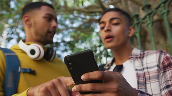 Two happy mixed race male friends standing, talking and using smartphone in the street