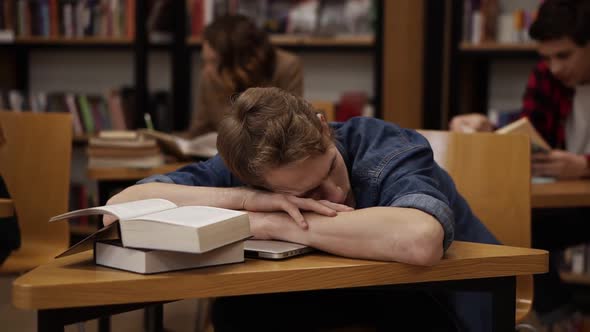 Young European Student in a College Library or Class Sleeping on Desk with Pile of Books on It