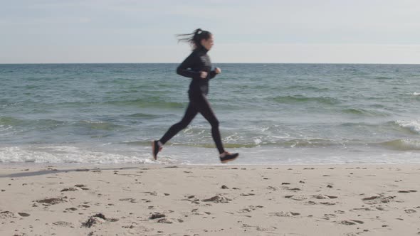 Jogging woman runs along beach. Static full body shot in slow motion. Bright blue sky and emerald gr