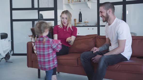 Back View of Little Girl Giving Gift Box To Smiling Mother Sitting with Man on Couch. Attentive