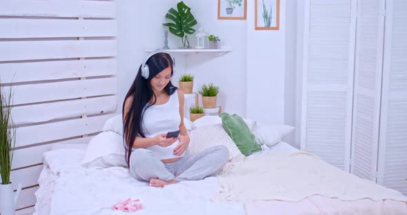 Pregnant Woman Is Resting in the Bedroom, Listening To Music on Headphones. Beautiful Pregnant Woman