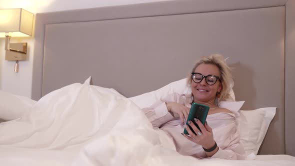 Cheerful Blonde Woman Is Lying in Bed in Hotel Room and Chatting By Video Call in Cellphone