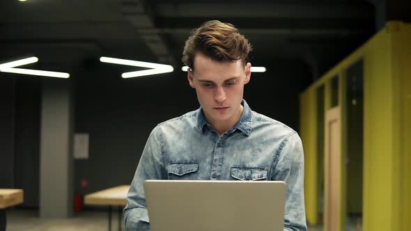 Young Attractive Guy in Denim Shirt with Laptop in His Hands is Walking Somewhere in Well Lit