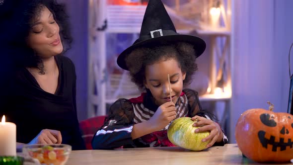 Little Girl and Woman African American in Festive Costume and Witch Hat Will Paint Pumpkin