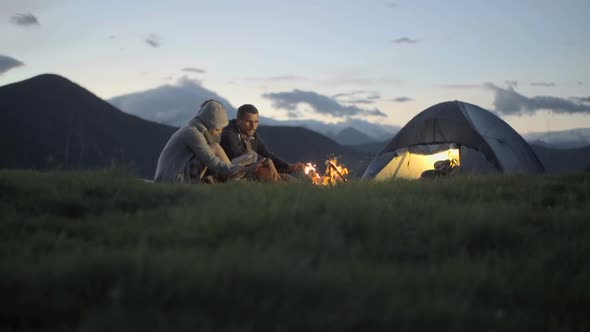 Group of Three Friends Warming with Camp Fire in Nature Mountain Outdoor Camping Scene at Night