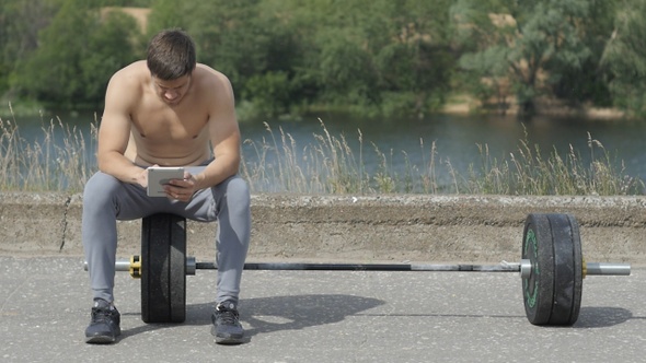 Young muscular man sitting after fitness training and using