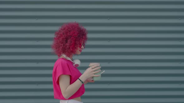 A Pink Curly Teenager is Walking and Using a Smartphone Outdoors alt