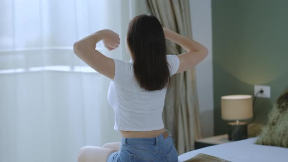 Back view of young woman raising her hands up and stretching arms sitting on the bed