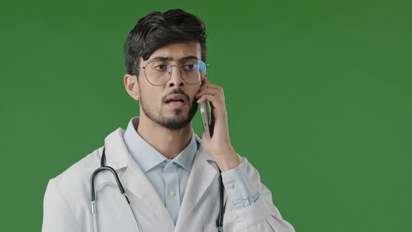 Excited Young Arabic Doctor Consulting Remotely Using Phone Serious Man Medic on Green Background in