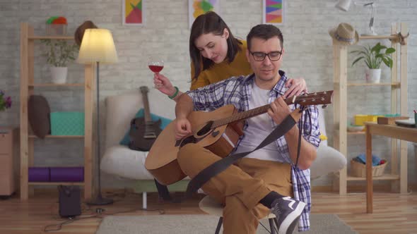 Young Man Musician with Glasses Playing Guitar He Is Approached By a Young Woman with a Glass of