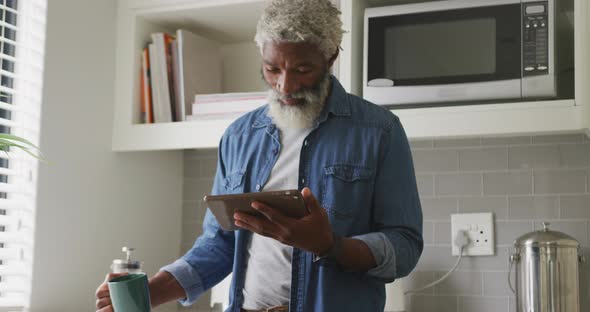 Video of happy african american senior man drinking coffee and using tablet