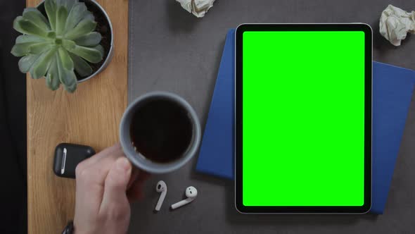 A Man Puts Cup of Coffee on the Desktop Next to a Tablet with a Green Screen