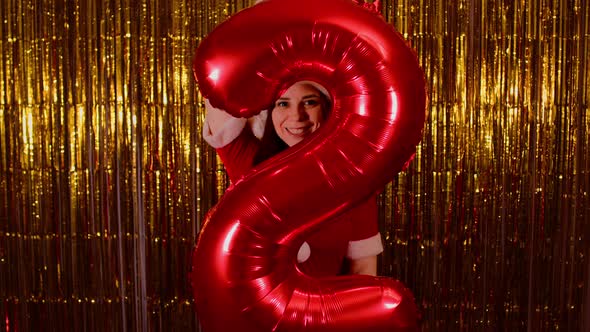 Woman in Santa Costume Holds Red Figure Two on Background of Shimmering Golden Tinsel