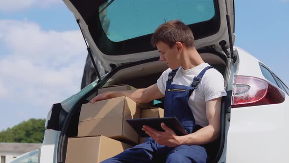 The Courier Sits on the Edge of the Open Trunk Filled with Boxes of Goods and Fills Out Documents