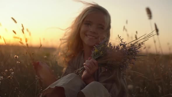 Portrait of Beautiful Little Girl with Blond Long Hair Sitting in Field Covered with Plants Holding