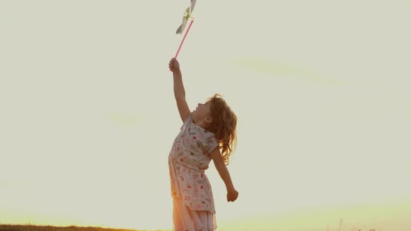Little Girl Standing On Nature At Sunset. Girl Holding Toy Windmill Moving In The Wind.