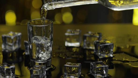 Barman Pour Frozen Vodka From Bottle Into Shot Glass. Ice Cubes Against Shiny Gold Party Background