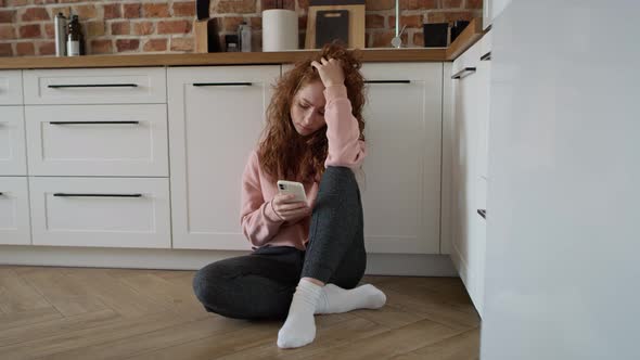 Depressed young caucasian woman sitting sad on floor in the kitchen.