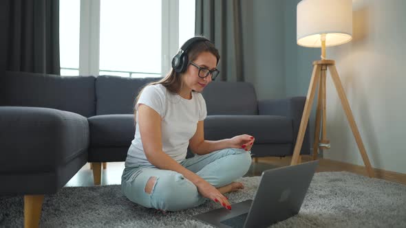 Casually Dressed Woman with Headphones is Sitting on Carpet with Laptop and Working in Cozy Room