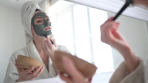 Beautiful Smiling Woman at Home in Bathrobe with a Towel Applying Face Clay Mask Against Acne