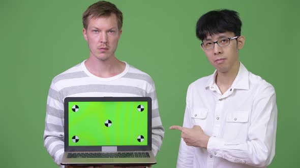 Two Young Multi-ethnic Businessmen Showing Laptop To the Camera While Pointing Finger