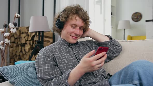 Guy with Curls in Headphones Lying on the Sofa in Contemporary Apartment and Enjoying Modern Music