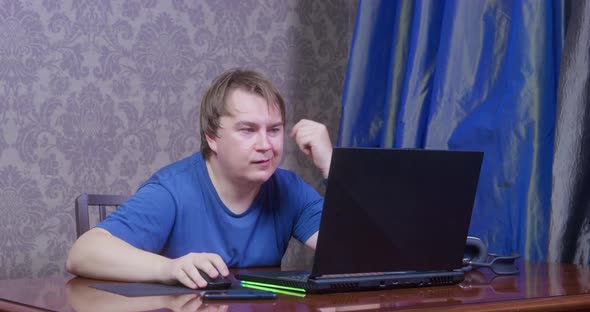 Young Bored Man in Blue Tshirt with a Silly Look Picks at His Ear and Scratches It Sits at Table in