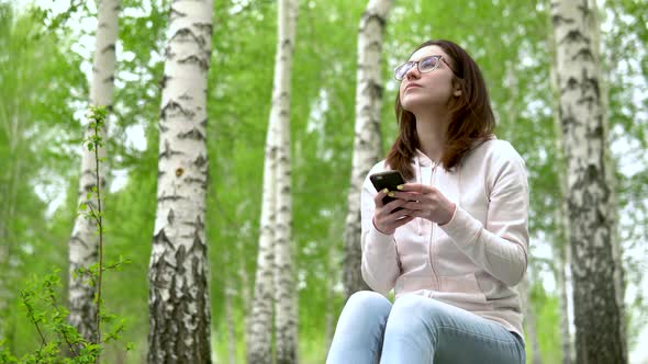 Young Woman in Nature with a Phone in Her Hands. A Girl Sits on a Stump in a Birch Forest and Leads